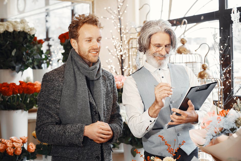 A florist with grey hair, beard, and glasses, dressed in a light grey vest and white shirt, is holding a tablet and selecting a floral arrangement in a shop filled with fresh flowers. Beside him is a