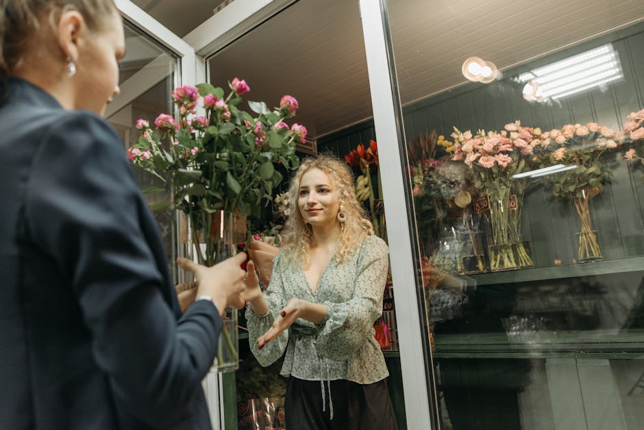 A florist at flowerscranham.co.uk handing over a bouquet of fresh pink roses wrapped in brown paper and tied with a pink ribbon to a customer inside a floral shop. The bouquet features vibrant pink ro