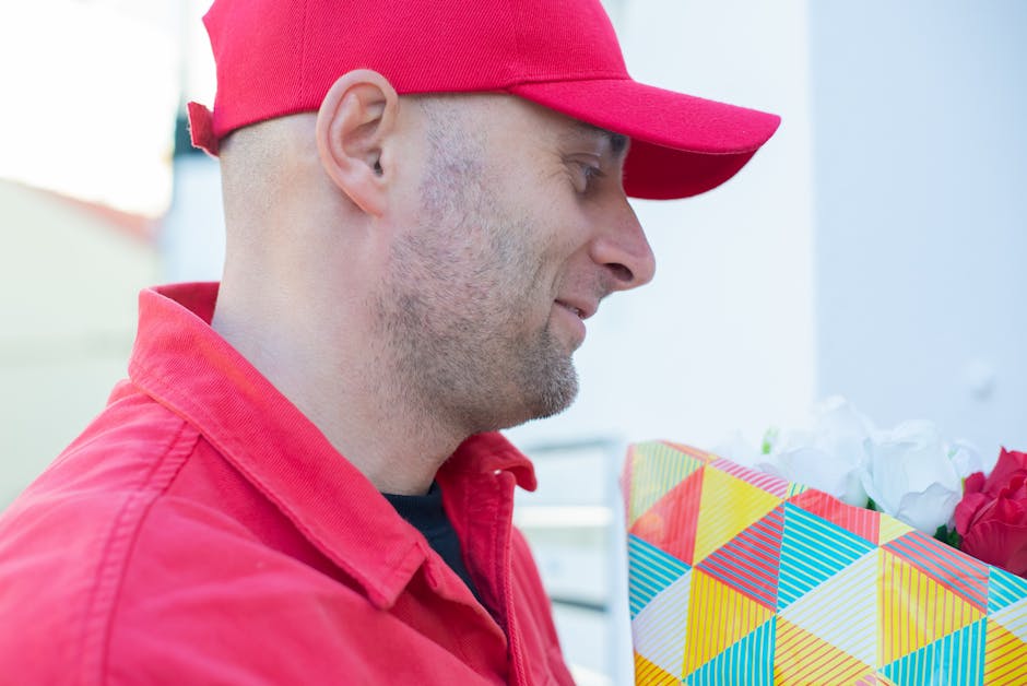 A delivery person dressed in a red cap and red jacket holding a floral gift box wrapped in colorful geometric-patterned paper with a grid design and crafted from sturdy material. The bouquet inside fe