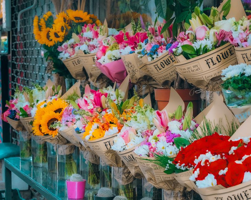 A display of colorful flower bouquets arranged on a glass shelf, featuring a variety of fresh flowers such as bright yellow sunflowers, pink lilies, white and pink roses, and clusters of blue and purp