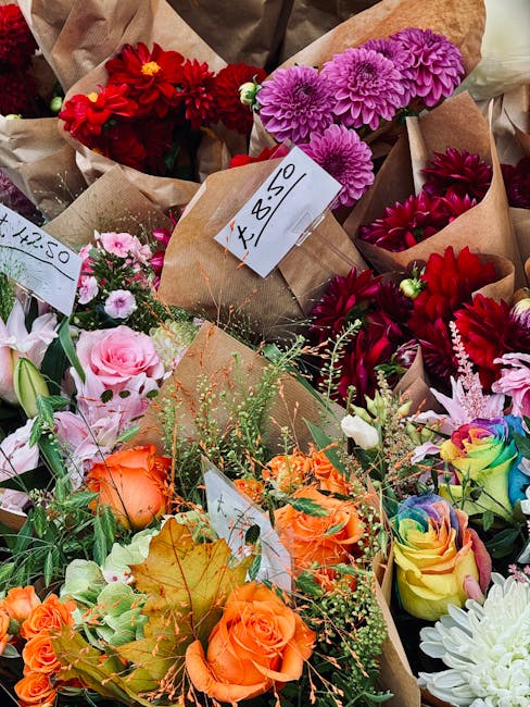 A vibrant assortment of fresh flowers including pink roses, orange roses, red dahlias, purple chrysanthemums, and multicolored roses with rainbow hues. The flowers are arranged in hand-tied bouquets,