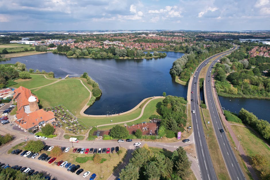 An aerial view showing a scenic, tranquil lake surrounded by lush green trees and grassy areas, with a pathway curving along the water's edge. To the left, there is a charming building with a red-tile